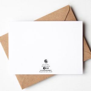 May include: A white greeting card with a brown envelope. The card is blank, with a small logo and text at the bottom. The envelope is a light brown color and partially visible behind the card.