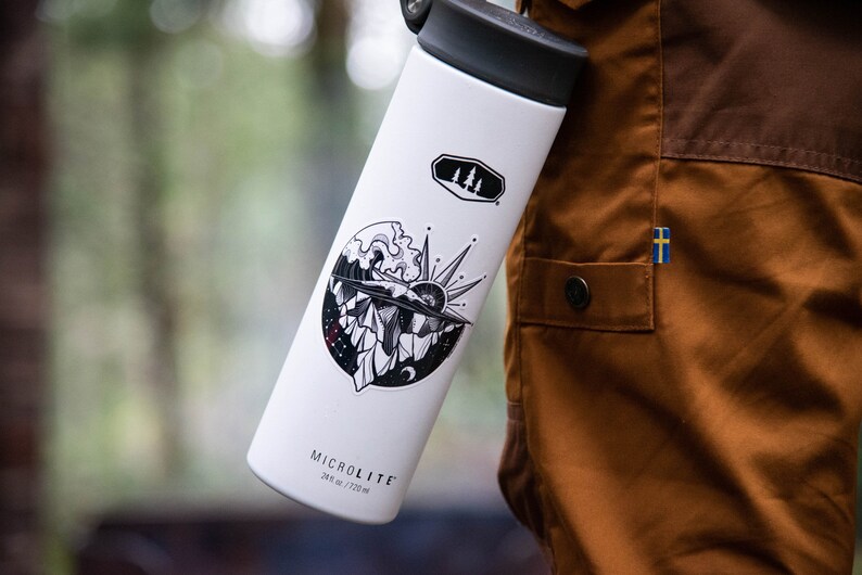 May include: White insulated water bottle with a black lid and a black and white sticker featuring a mountain range, a sun, and a wave. The sticker also includes a small moon and stars. The bottle has the text "MICROLITE 24 oz / 720 ml" printed on the bottom.