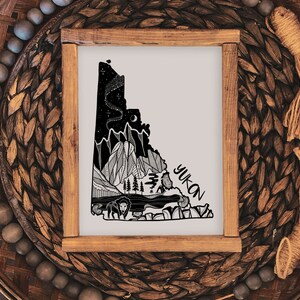 May include: A framed art print featuring a black and white illustration of the Yukon territory. The artwork depicts mountains, a lake, trees, and wildlife, with the word "Yukon" in a handwritten font. The frame is made of light brown wood.
