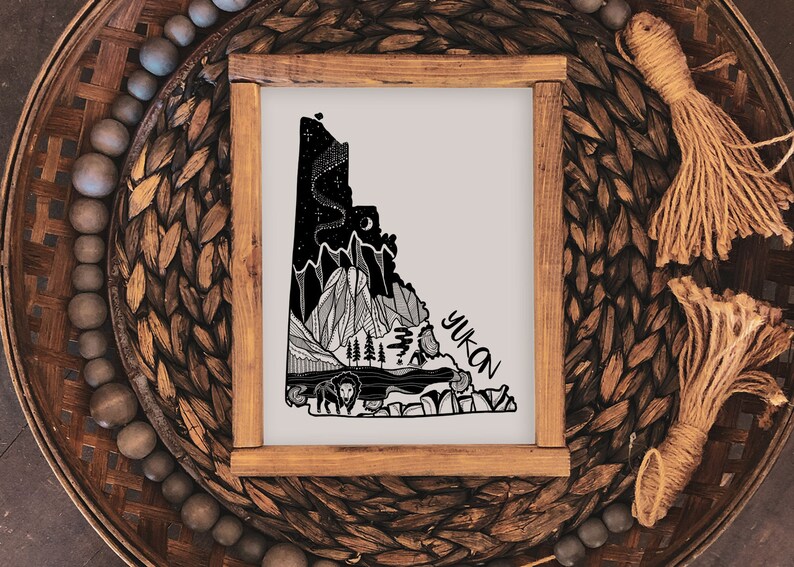 May include: A framed art print featuring a black and white illustration of the Yukon territory. The artwork depicts mountains, a lake, trees, and wildlife, with the word "Yukon" in a handwritten font. The frame is made of light brown wood.