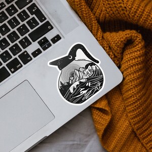 May include: Black and white sticker of a coffee pot with a mountain scene inside. The sticker is on a silver laptop.
