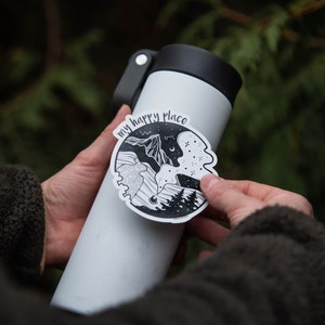 May include: A white water bottle with a black lid and a black and white sticker that says "my happy place" with a mountain and forest scene.