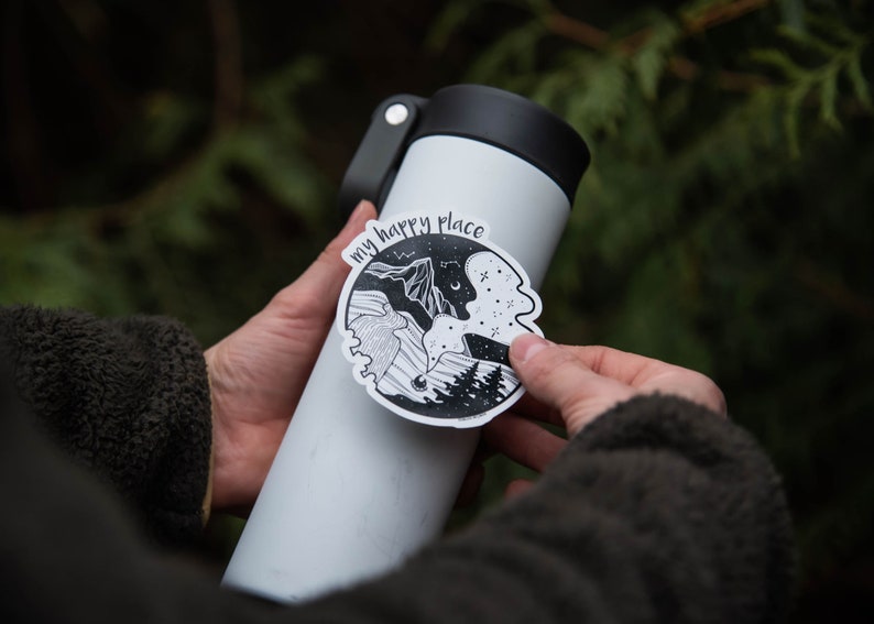 May include: A white water bottle with a black lid and a black and white sticker that says "my happy place" with a mountain and forest scene.