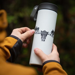 Puede incluir: Una taza de viaje blanca con una tapa negra y una pegatina en blanco y negro de un alce con un sombrero y gafas. La pegatina se est&aacute; aplicando a la taza.