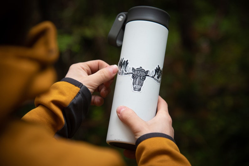 Puede incluir: Una taza de viaje blanca con una tapa negra y una pegatina en blanco y negro de un alce con un sombrero y gafas. La pegatina se est&aacute; aplicando a la taza.