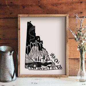 May include: A framed art print featuring a black and white illustration of the Yukon territory. The artwork depicts mountains, trees, a bison, and a night sky with the word "Yukon" in a stylized font. The print is displayed on a wooden surface.