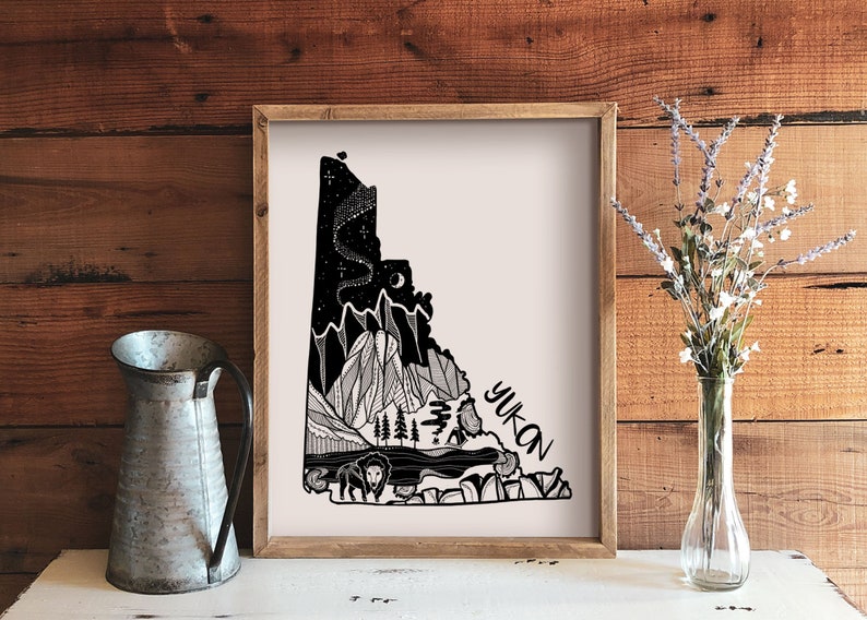 May include: A framed art print featuring a black and white illustration of the Yukon territory. The artwork depicts mountains, trees, a bison, and a night sky with the word "Yukon" in a stylized font. The print is displayed on a wooden surface.