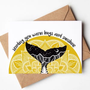 May include: A white greeting card featuring a black whale tail design, a yellow mandala, and the text "sending you warm hugs and sunshine." The card rests on a brown envelope.