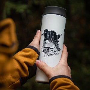 Pode incluir: Uma caneca de viagem branca com tampa preta e um autocolante com um gr&aacute;fico a preto e branco da prov&iacute;ncia de New Brunswick. O autocolante inclui o texto "NEW BRUNSWICK". A caneca &eacute; segurada por uma pessoa que usa um casaco amarelo.