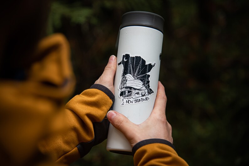 Pode incluir: Uma caneca de viagem branca com tampa preta e um autocolante com um gr&aacute;fico a preto e branco da prov&iacute;ncia de New Brunswick. O autocolante inclui o texto "NEW BRUNSWICK". A caneca &eacute; segurada por uma pessoa que usa um casaco amarelo.