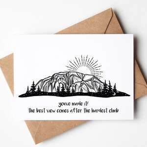 May include: A white greeting card with a black and white mountain illustration, a rising sun, and pine trees. The card includes the text "you've made it! the best view comes after the hardest climb." The card is placed on a brown envelope.