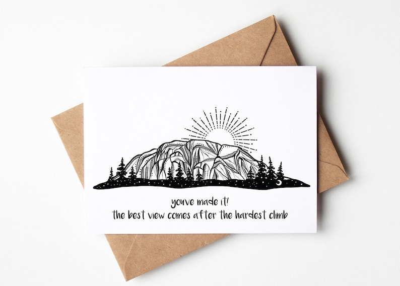 May include: A white greeting card with a black and white mountain illustration, a rising sun, and pine trees. The card includes the text "you've made it! the best view comes after the hardest climb." The card is placed on a brown envelope.