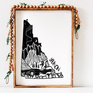 May include: Black and white art print of the Yukon territory, framed in a wooden frame. The artwork features a detailed illustration of mountains, trees, a bison, and the text "YUKON". The night sky is depicted with stars and a crescent moon.