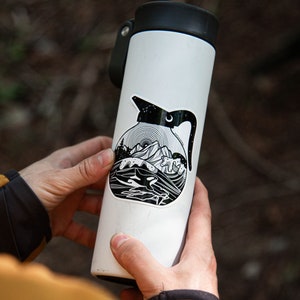 May include: A white water bottle with a black and white sticker of a coffee pot with a mountain scene and a whale in the water. The sticker has a black outline and a white interior.