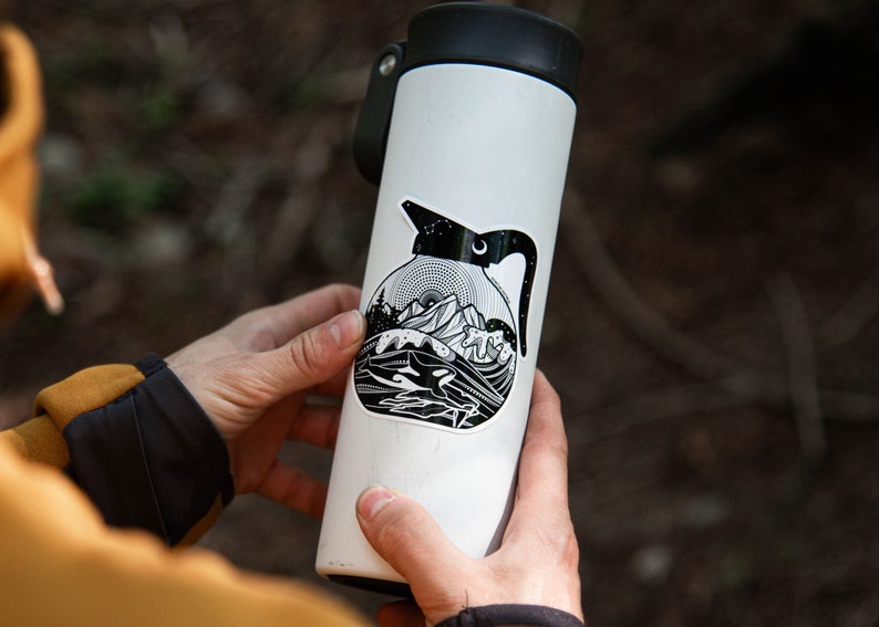 May include: A white water bottle with a black and white sticker of a coffee pot with a mountain scene and a whale in the water. The sticker has a black outline and a white interior.