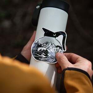 May include: A black and white sticker of a coffee pot with a mountain scene inside. The sticker is being applied to a white water bottle.