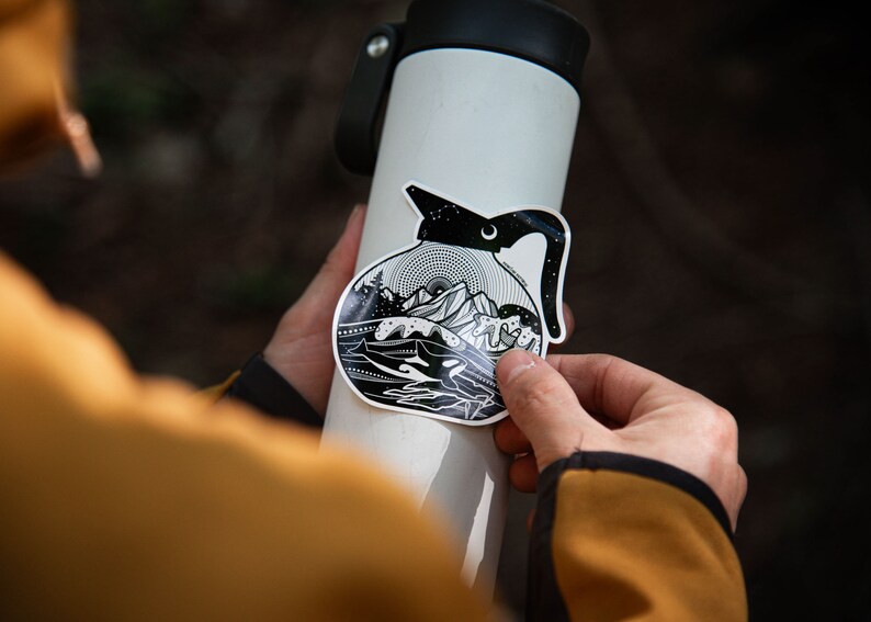 May include: A black and white sticker of a coffee pot with a mountain scene inside. The sticker is being applied to a white water bottle.