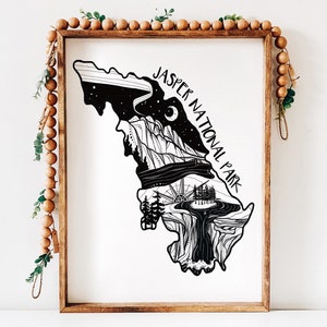May include: Black and white art print of Jasper National Park in a wooden frame. The artwork depicts a stylized map of the park with mountains, trees, and a night sky with the text "JASPER NATIONAL PARK". Decorative wooden beads and greenery surround the frame.