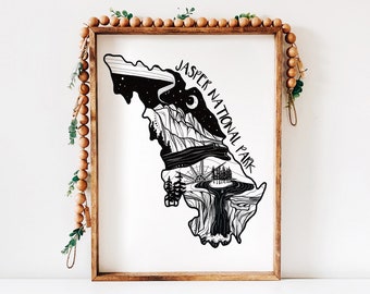 May include: Black and white art print of Jasper National Park in a wooden frame. The artwork depicts a stylized map of the park with mountains, trees, and a night sky with the text "JASPER NATIONAL PARK". Decorative wooden beads and greenery surround the frame.