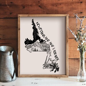 May include: A framed print with a black and white illustrated map of Newfoundland and Labrador. The artwork includes mountain and forest details, with the text "NEWFOUNDLAND AND LABRADOR". A galvanized metal jug and a vase of flowers are in the foreground.