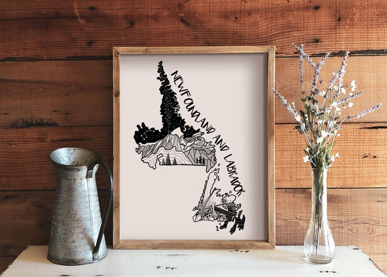 May include: A framed print with a black and white illustrated map of Newfoundland and Labrador. The artwork includes mountain and forest details, with the text "NEWFOUNDLAND AND LABRADOR". A galvanized metal jug and a vase of flowers are in the foreground.