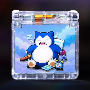 May include: A clear, transparent handheld gaming console shell featuring a pixel art graphic of a sleeping Snorlax character on a blue sky background with clouds, a small alarm clock, a book, and a flute, with visible internal electronic components.