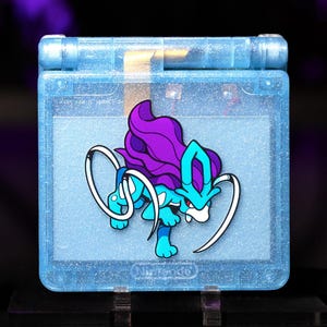 Op de afbeelding: Een doorschijnende lichtblauwe glitter handheld spelcomputer met een afbeelding van het personage Suicune in paarse en blauwgroene kleuren. Het apparaat heeft een vierkante vorm met een scharnier aan de bovenkant en het Nintendo-logo onderaan.