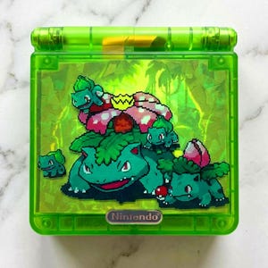 May include: A green Nintendo Game Boy Advance SP. The screen features a colourful graphic of the Pokémon character Venusaur and its pre-evolutions, set against a green, leafy background. The Nintendo logo is at the bottom.