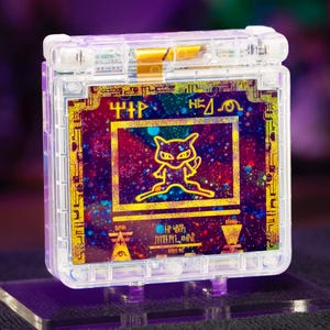 May include: A clear, transparent handheld gaming console shell featuring a vibrant, multi-coloured galaxy-themed insert. The centre displays a yellow outline of a Mew character, surrounded by yellow geometric symbols and text on a dark, speckled background.