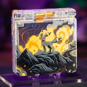 以下が含まれることがあります： 透明なプラスチック製の携帯ゲーム機。黄色い輪を持つ黒い生き物が岩山の頂上に立ち、渦巻く黄色い雲、オレンジ色の星、暗い夜空を背景にしたグラフィックが施されており、内部の部品が透けて見えています。