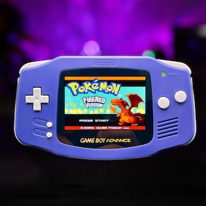 Puede incluir: Una consola portátil Nintendo Game Boy Advance de color morado con una cruceta blanca, botones A y B, y botones Start y Select. La pantalla muestra la pantalla de título del videojuego Pokemon FireRed Version con una ilustración de Charizard.