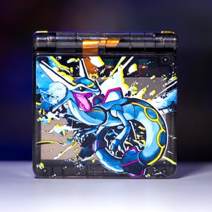 Könnte beinhalten: Eine transparente Handheld-Spielkonsole mit einer lebendigen Illustration eines blau-weißen drachenartigen Wesens mit gelben Akzenten. Die Konsole hat ein schwarzes Scharnier und das Nintendo-Logo ist sichtbar.