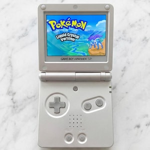 May include: A silver Game Boy Advance SP handheld gaming console with a flip-top screen. The screen displays the title screen for Pokémon Liquid Crystal Version, featuring a blue creature. The device includes a D-pad, A and B buttons, and start buttons.