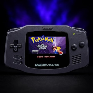 May include: A dark grey Game Boy Advance console, displaying the Pokémon Ash Gray Version game. The screen shows the game's title and characters. The console has a Nintendo logo and control buttons.