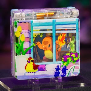 May include: A transparent purple handheld gaming console featuring a custom graphic on the lid. The design depicts a Charmander character inside a window, a Pidgey bird, yellow tulips, and purple flowers. The Nintendo logo is visible at the bottom.
