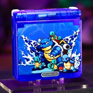 Peut inclure: Une console de jeu portable bleu translucide avec un graphique en pixel art représentant Blastoise, Squirtle et Wartortle entourés d'éclaboussures d'eau. Le logo Nintendo est imprimé en argent sur le bas de la façade de l'appareil.