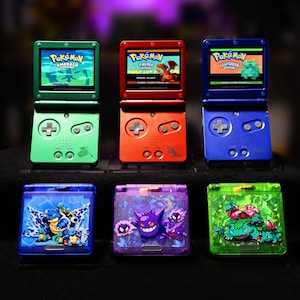 May include: Six Game Boy Advance SP handheld consoles. The top row features green, red, and blue units displaying Pokémon Emerald, FireRed, and LeafGreen. The bottom row shows three translucent units in blue, purple, and green with character graphics.
