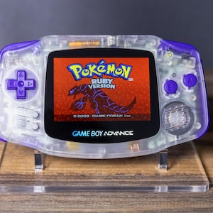 Könnte beinhalten: Ein transparentes, lila Game Boy Advance Handheld-Konsolen-Gerät, das das Spiel "Pokémon Ruby Version" anzeigt. Die Konsole hat lila Knöpfe und ein Steuerkreuz. Der Bildschirm zeigt einen roten Hintergrund mit einer Pokémon-Grafik. Der Text "Game Boy Advance" ist sichtbar.
