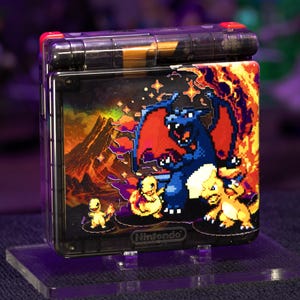 以下が含まれることがあります： 蓋にカスタムグラフィックが施された半透明の黒い携帯ゲーム機。アートワークには、炎のある火山風景を背景に、ピクセル化された青いリザードン、ヒトカゲ、リザードが描かれています。下部にはNintendoのロゴが刻印されています。
