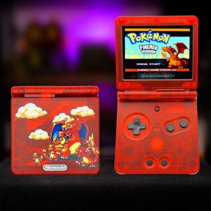 May include: A translucent red Game Boy Advance SP handheld console. One unit is closed, displaying a graphic of Charizard and Charmander with the Nintendo logo. The other unit is open, showing the Pokémon FireRed Version title screen.
