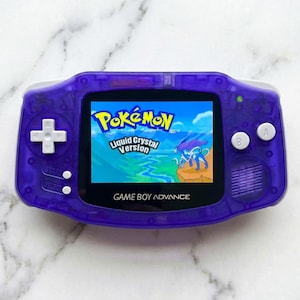 May include: A purple Game Boy Advance handheld console displaying the "Pokémon Liquid Crystal Version" game. The screen shows a vibrant landscape with the Pokémon logo. The console has white buttons and a directional pad.