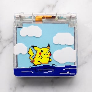 Puede incluir: Carcasa transparente para Game Boy Advance SP con un diseño personalizado. El diseño presenta un personaje Pikachu pixelado sobre un fondo de cielo azul con nubes blancas. El diseño es visible a través de la carcasa transparente.