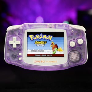 May include: A translucent purple Game Boy Advance handheld console featuring white buttons and a screen displaying the title screen for Pokémon ShinyGold Version, which includes the text Press Start and 2006-2008 by Zel with a bird graphic.
