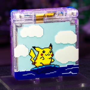 May include: A handheld video game console with a transparent purple plastic shell. The front cover features a pixel art graphic of a yellow Pikachu character surfing on a blue wave against a light blue sky with white pixelated clouds.