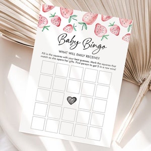 May include: A white Baby Bingo game card with a strawberry design at the top. The card has a grid of squares and the text "Baby Bingo" and "What will Emily receive?" is printed on the card. The card is on a white plate.