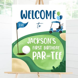 May include: Welcome to Jackson's 1st Birthday Par-Tee sign with a green golf course, a golf cart, a golf ball, and blue and green balloons.