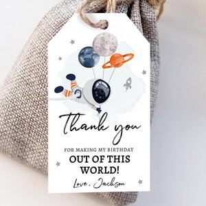 May include: A white thank you tag with a watercolor illustration of an astronaut holding onto balloons shaped like planets. The text on the tag reads "Thank you for making my birthday out of this world! Love, Jackson".