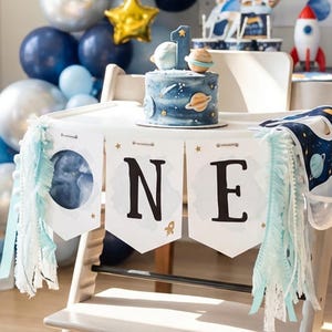 Op de afbeelding: Een kinderstoel versierd met een banner in ruimtethema waarop in zwarte letters ONE staat. De banner heeft lichtblauwe franjes aan de zijkanten. Een kleine ronde taart met ruimtedecoraties staat op het blad van de kinderstoel.
