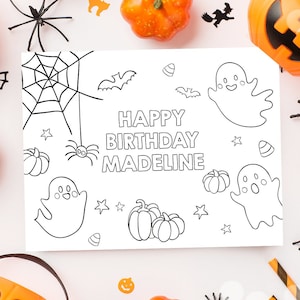 May include: A Halloween-themed birthday card with the text "Happy Birthday Madeline" surrounded by pumpkins, spiders, bats, and ghosts. The card is black and white, with orange and black decorations scattered around it.