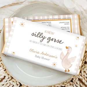May include: A rectangular chocolate bar wrapper featuring a white background with a light beige gingham border. The text reads A NEW silly goose IS ABOUT TO BE ON THE LOOSE! Olivia Anderson's Baby Shower. A watercolor illustration of a goose and daisies.